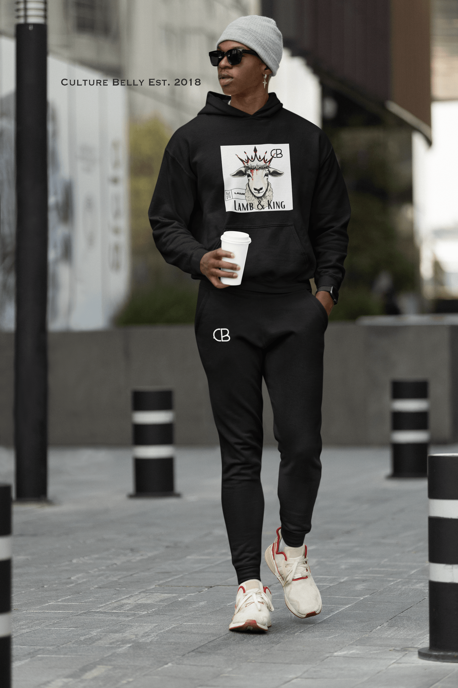Stylish man wearing the Lamb & King Sweatsuit bundle by Culture Belly's Always Faithful Faithwear collection, featuring a bold hoodie and sweatpants with the CB logo, walking confidently in an urban setting while holding a coffee cup.