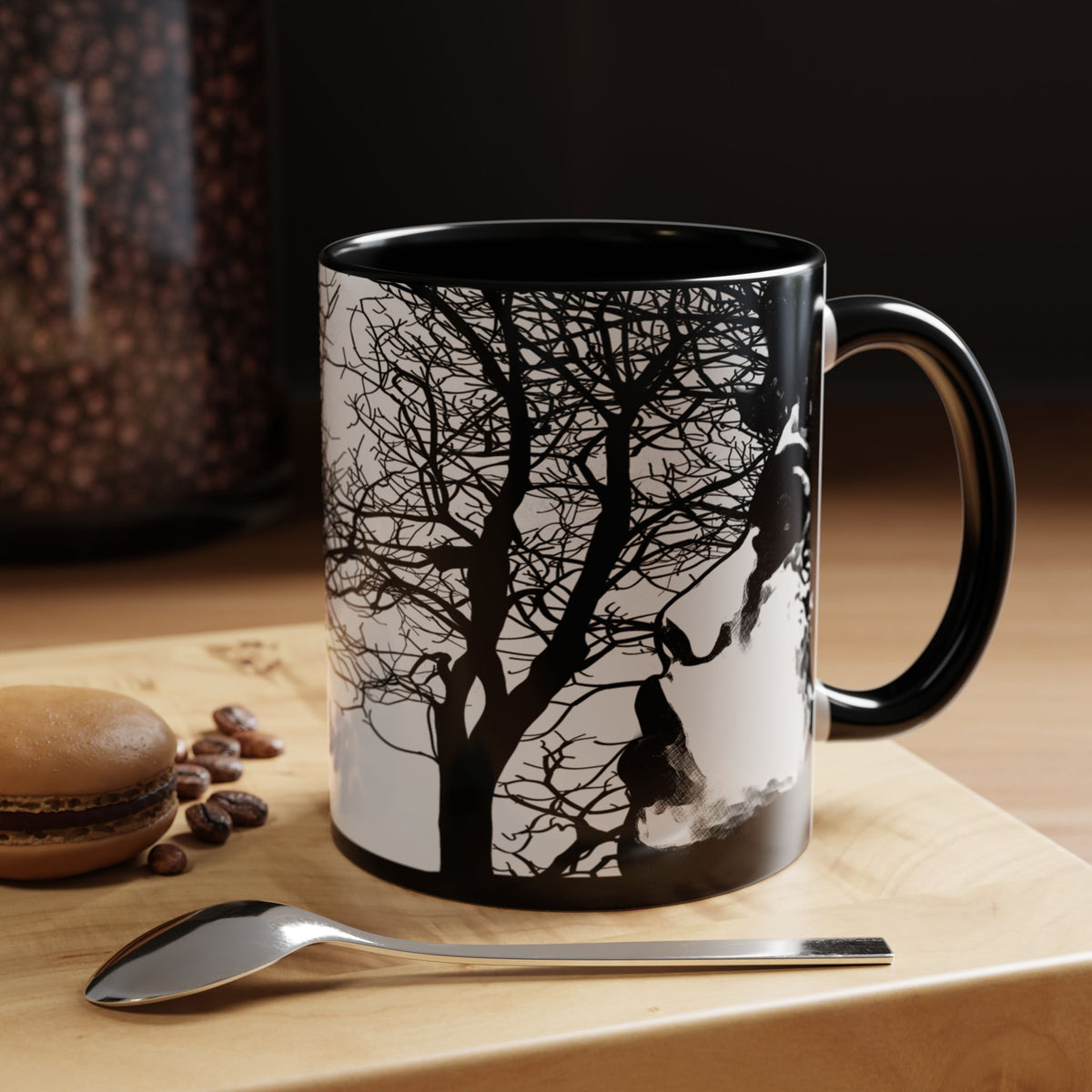 Black Through The Wilderness ceramic coffee mug displayed on a wooden tabletop with coffee beans and a pastry, featuring a black and white forest silhouette with a contemplative face illustration wrapping around the mug.
